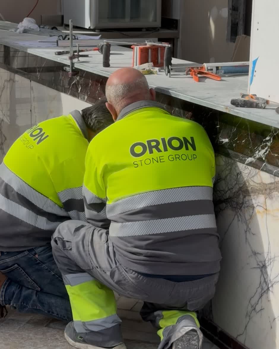 Dos operarios de Orion agachados, ajustando el frente de mármol de una barra en obra.